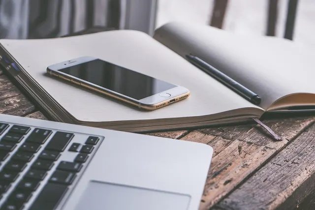 Smartphone resting on an open blank notebook with a pen, beside a laptop keyboard on a weathered wooden desk