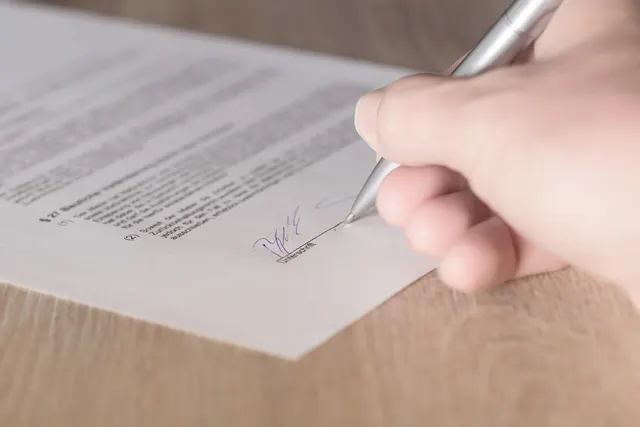 Close-up of a hand signing a printed contract with a silver pen on a wooden desk