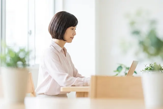 Woman with a bob haircut in a light pink blouse working on a laptop at a wooden table in a bright room with potted plants