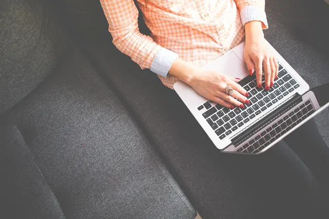 Overhead view of a woman in a peach gingham shirt with red nails typing on a silver laptop on a gray couch