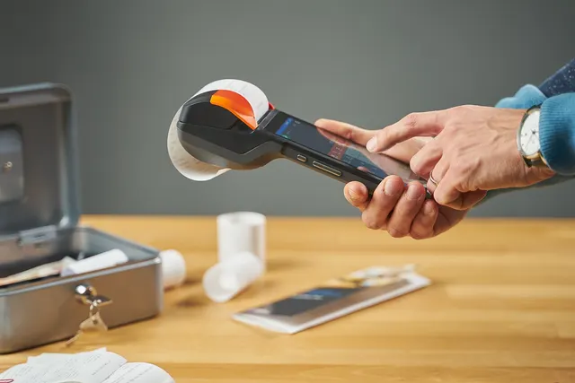 Hands holding a handheld POS terminal printing a receipt, beside an open cash box and paper rolls on a wooden counter