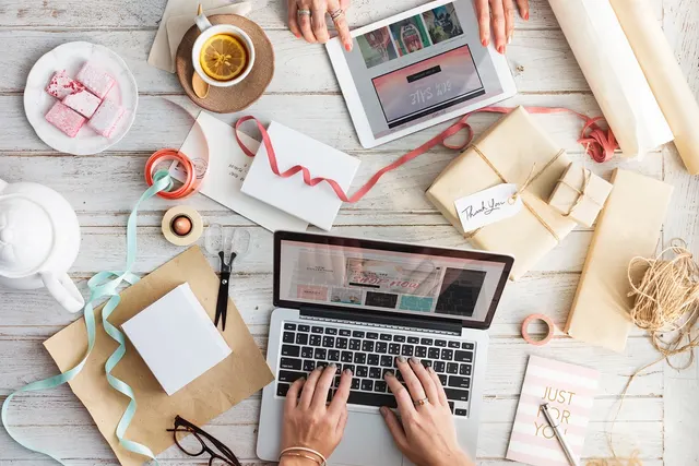 Overhead view of hands typing on a laptop showing an online shop, beside a tablet, wrapped gift boxes, ribbon and tea