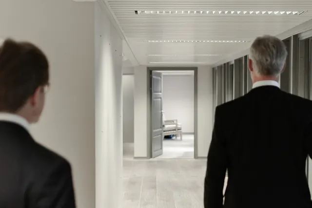 Two men in dark suits, seen from behind, walk down a bright office corridor toward an open door and a chair