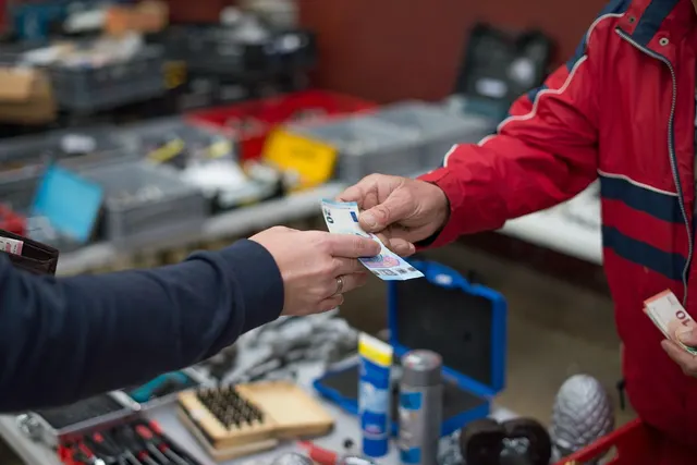 Customer handing euro banknotes to a vendor in a red jacket across a market stall table of tools and hardware
