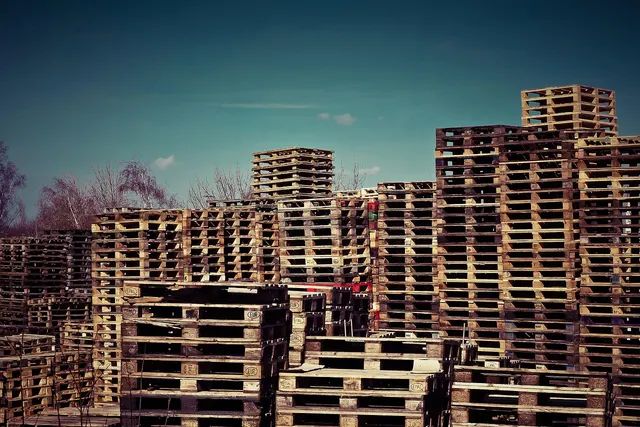Tall stacks of wooden shipping pallets piled high in an outdoor storage yard under a blue sky with bare trees