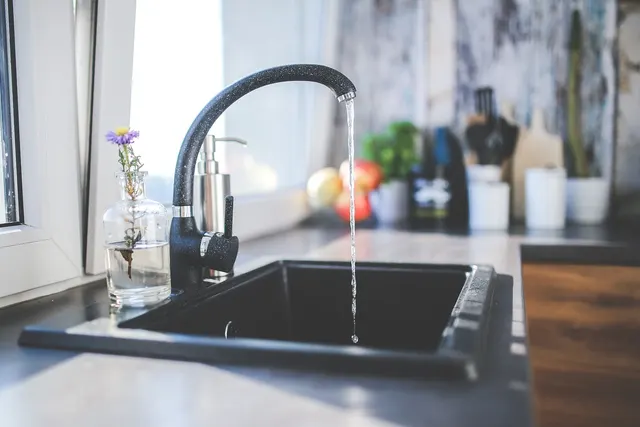 Black kitchen faucet running water into a dark sink beside a soap dispenser and a small flower in a glass vase