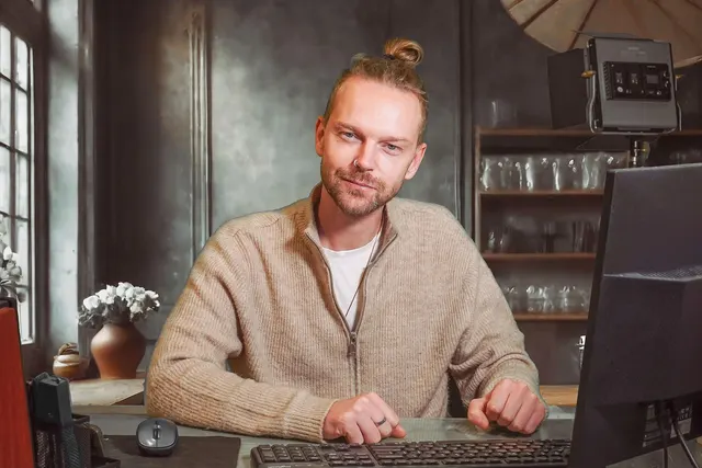 Smiling man in a beige sweater at a desk with keyboard, mouse and monitor, studio light and umbrella behind him