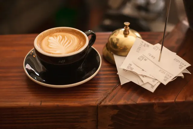 Latte with leaf art in a black cup beside a brass service bell and a stack of receipts on a spike on a wooden cafe counter