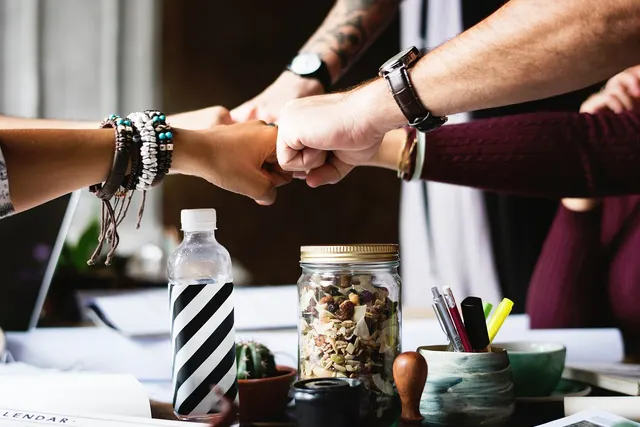 Team members bump fists over a cluttered office desk with a laptop, pens, a snack jar and a striped water bottle