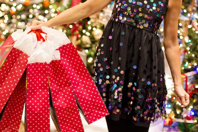 Woman in a sequined dress holding red polka-dot shopping bags in front of a lit Christmas tree