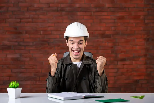 Excited contractor in a white hard hat pumping his fists at a desk with an open notebook against a brick wall