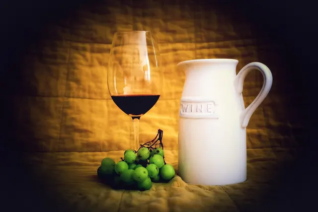Glass of red wine beside a white ceramic pitcher embossed with WINE and a bunch of green grapes on a gold cloth