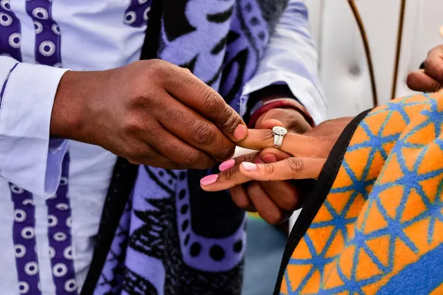Close-up of a man sliding a diamond ring onto a woman's finger, both wearing traditional patterned blankets