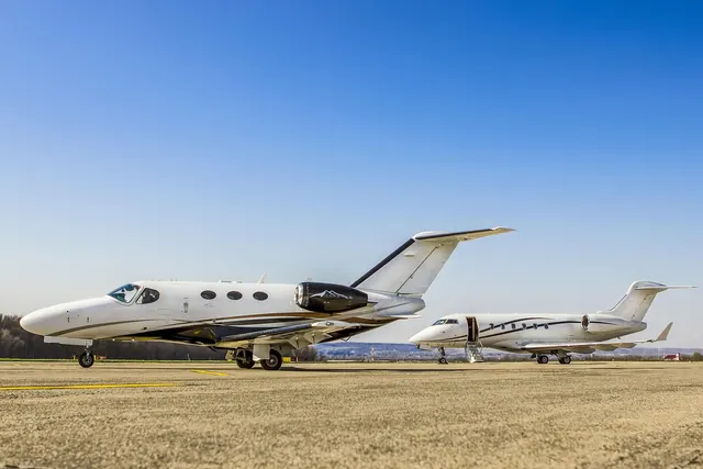 Two white private jets parked on a sunny airport apron under a clear blue sky, the nearer one with its door closed
