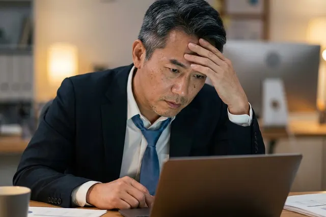 Stressed businessman in a suit holding his forehead while looking at a laptop in a dimly lit office at night