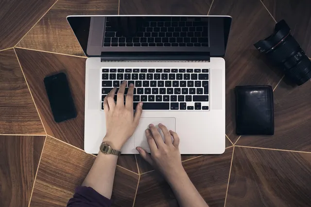Overhead view of hands typing on a laptop on a wooden desk beside a smartphone, black wallet and camera lens
