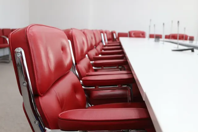 Row of red leather office chairs along a long white conference table with microphones in an empty boardroom