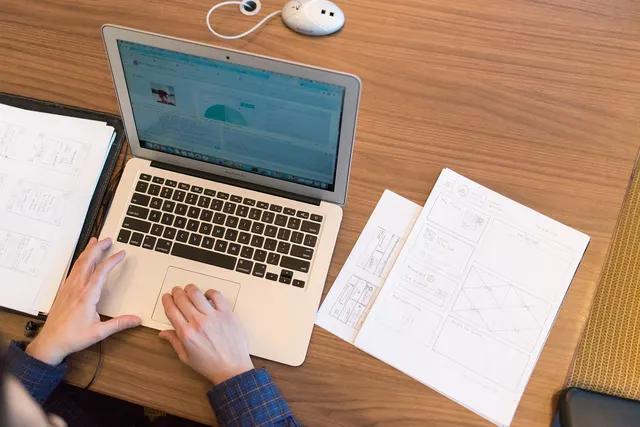 Overhead view of hands on a laptop showing an analytics dashboard, with printed website wireframe sketches on a wooden desk
