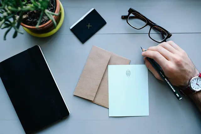 Overhead view of a hand holding a pencil over a blank notecard and kraft envelope on a gray desk with glasses and notebook